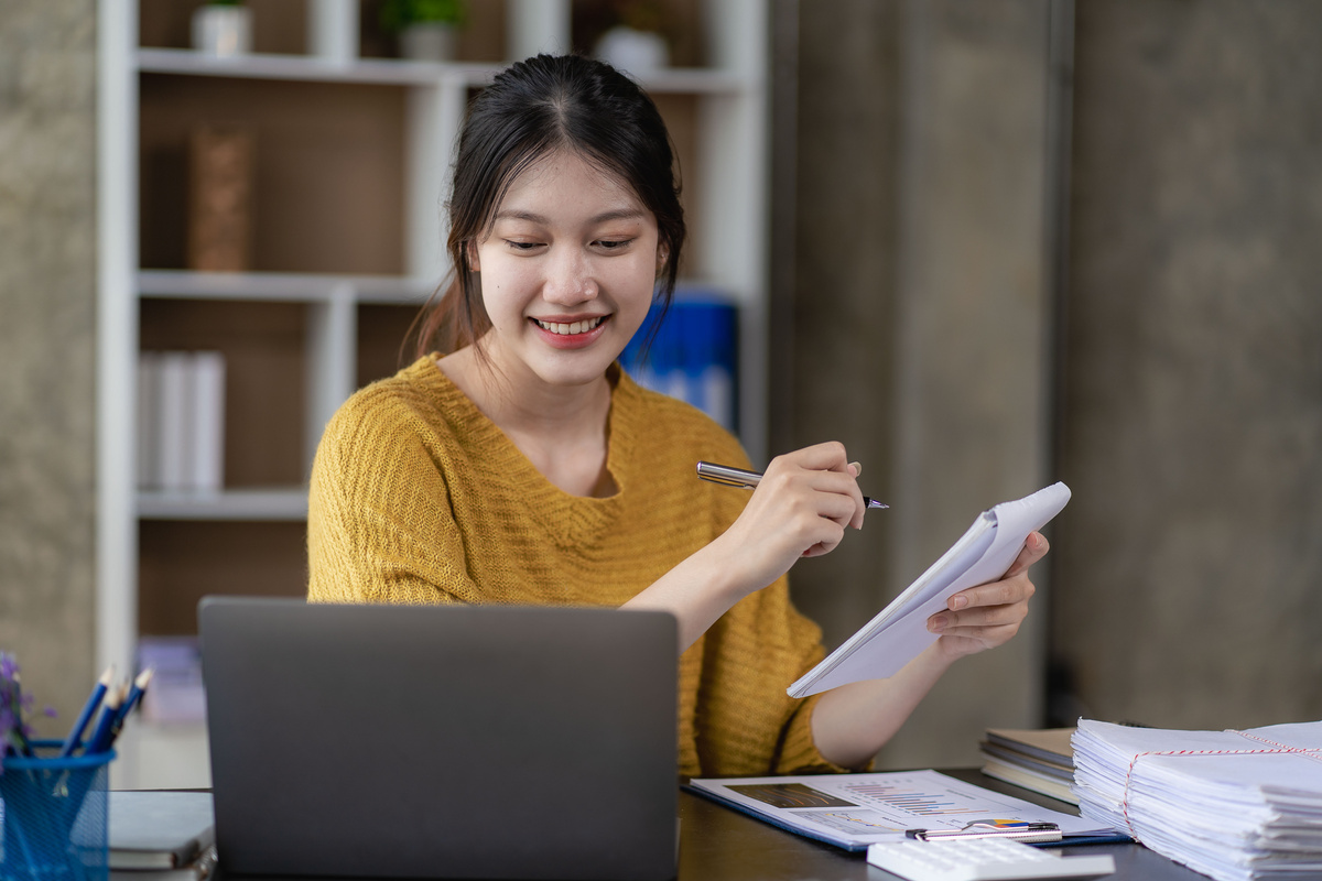 Asian businesswoman using smartphone and laptop writing on charts and graphs showing results Beautiful female accountant using calculator to calculate savings at home concept of working at home
